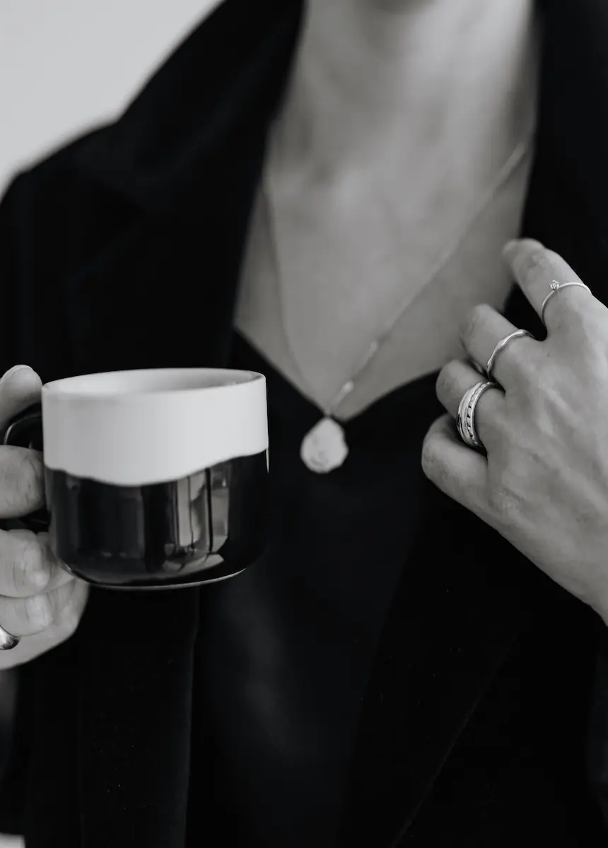 FAQ-Webdesign-Fragen-und-Antworten-zu-webdesign Nahaufnahme einer Person mit schwarzer Kleidung, die eine Tasse hält und mehrere Ringe sowie eine Kette mit Anhänger trägt.